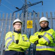 Two electricity North West engineers arms-crossed in front of fence, electricity pylon, and a danger of death sign