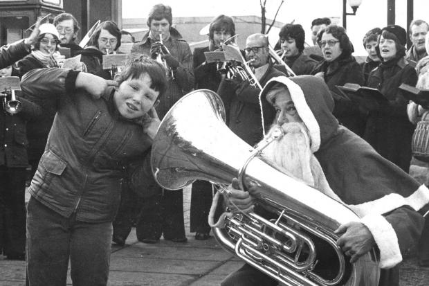 Mark Platt (9) finds the music a bit loud as Father Christmas starts off the carol singing at Harwood precinct.  The singers are the Harwood Methodist choir accompanied by Eagley Band. Photo by Bolton Evening News, December 18 1976..